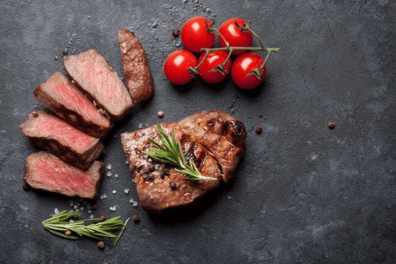 Sliced NY strip steak with rosemary, peppercorns, and cherry tomatoes on a dark surface